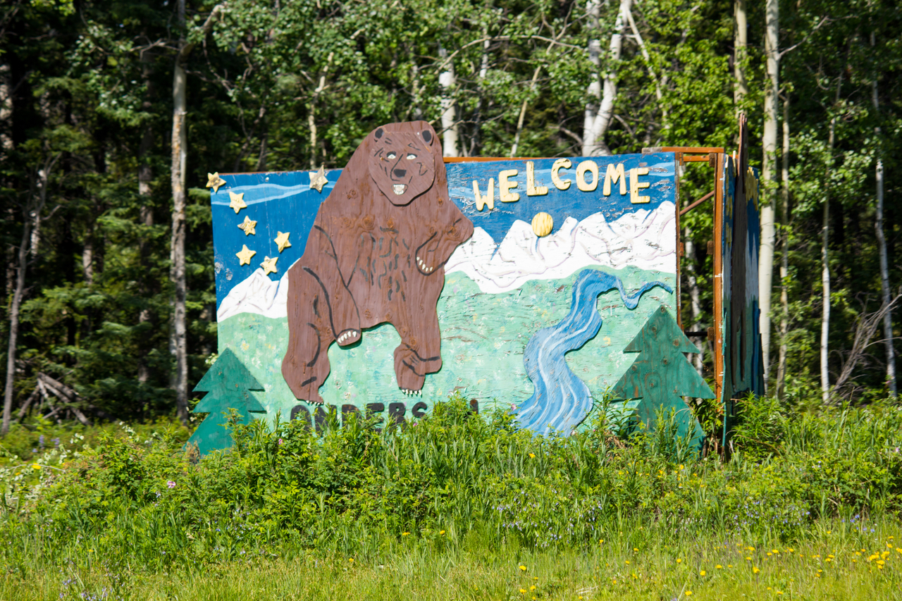 A wooden welcome sign for Anderson featuring a bear, mountains, river, forest and stars from the Alaska state flag.