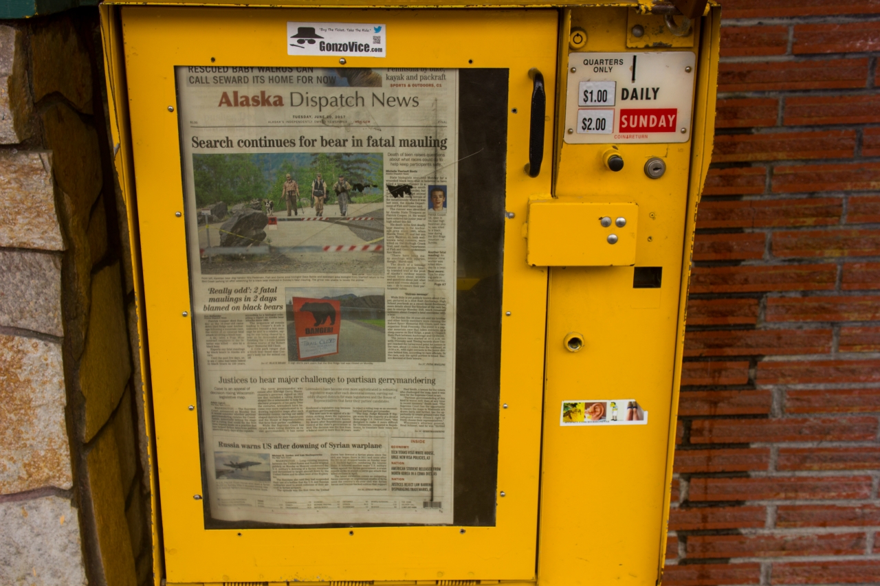 A yellow newspaper machine dispensing Alaska Dispatch News along the brick wall fronting Thorn's Showcase Lounge.