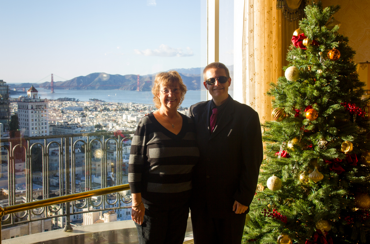 Carol Nichelson and David July pose with the Golden Gate Bridge (1937) beyond and a Christmas tree in the Crown Room lobby on 24F in the Tower Building (1961) of The Fairmont Hotel (1907).