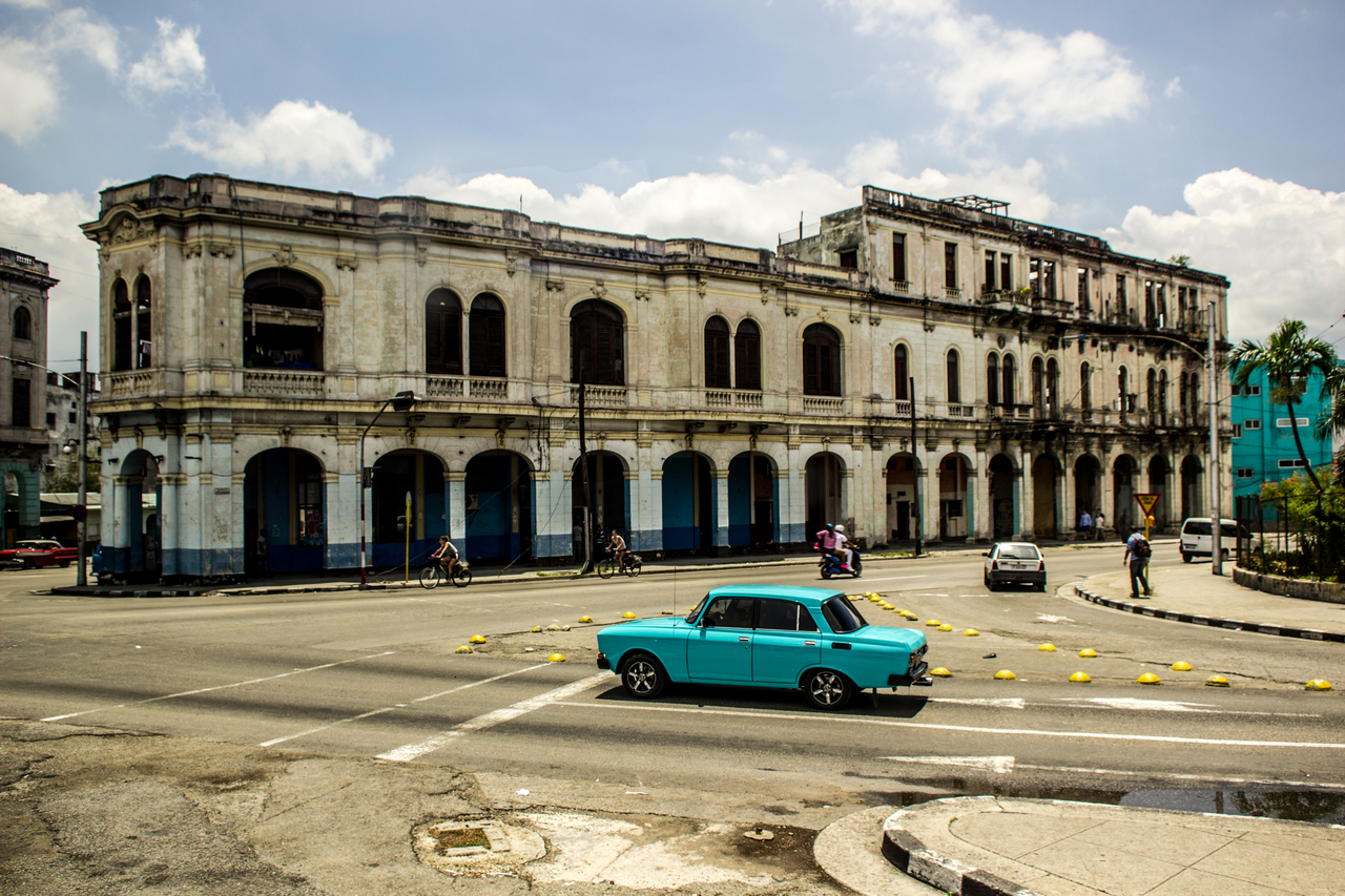 A sky blue Soviet Moskvitch 2140 (1976–1988) stopped at a five-way intersection in front of a building with arches in Havana, Cuba.