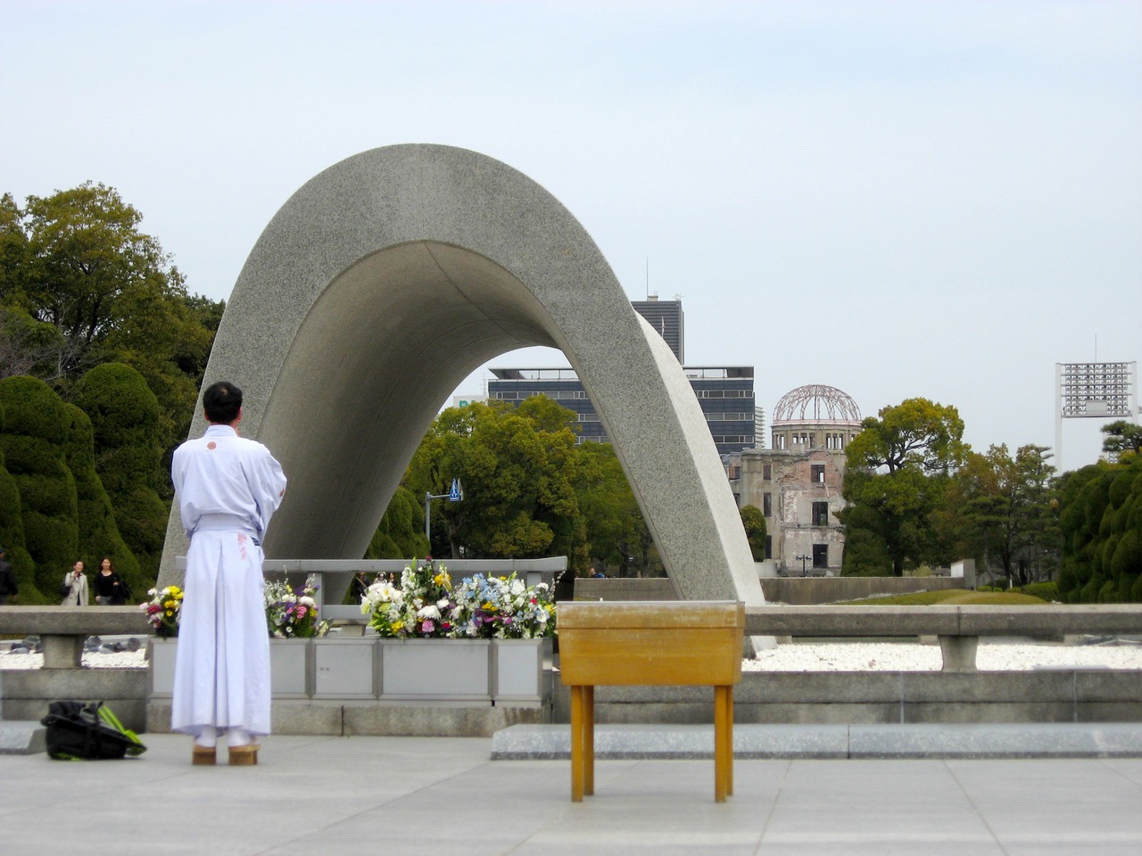 A man in a robe stands in remembrance at the Cenotaph for the A-Bomb Victims (Memorial Monument for Hiroshima, City of Peace) with the Genbaku Dome behind.