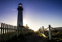 The sun starting to set behind Pigeon Point Lighthouse (1871) casts light over the watch house, fence and path.