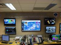Sign, NWS logo and wall-mounted displays in the Tropical Analysis and Forecast Branch unit of the National Hurricane Center.