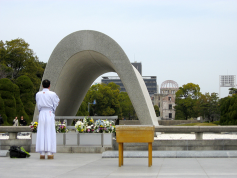Photo Credit: David July — A man in a robe stands in remembrance at the Cenotaph for the A-Bomb Victims (Memorial Monument for Hiroshima, City of Peace) with the Genbaku Dome behind, Hiroshima, Hiroshima Prefecture, Japan, 17 March 2008