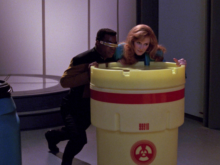 La Forge and Crusher push an overpack drum across the cargo bay floor