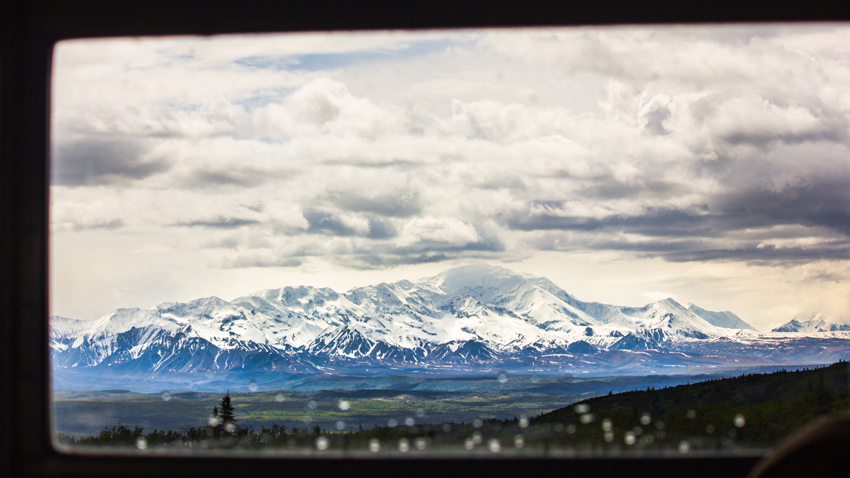 A vast area of wilderness leads to Alaska Range slopes and glaciers surrounding Denali, cloaked behind dense clouds, from through a starboard window aboard the Kantishna Experience tour bus.