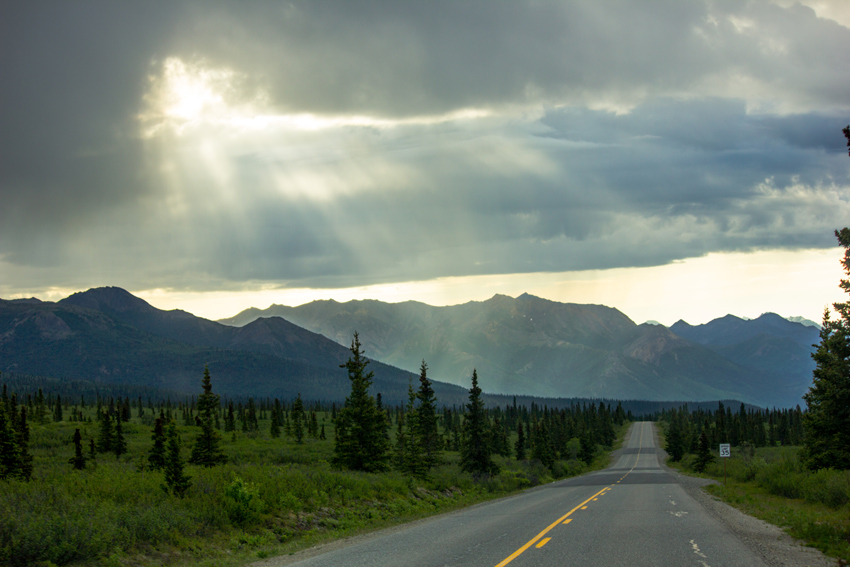 Sunbeams shining through clouds and extending across the sky above the forest and mountains in and around Denali National Park while driving east on Park Road.