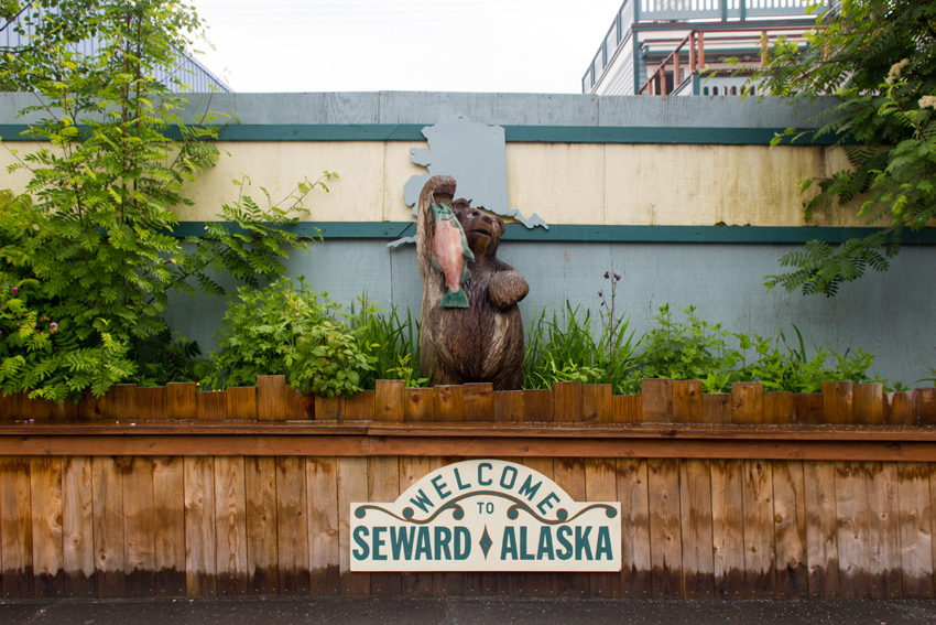 Wooden carving of a bear holding a fish at the 'Welcome To Seward Alaska' sign between storefronts on Fourth Avenue